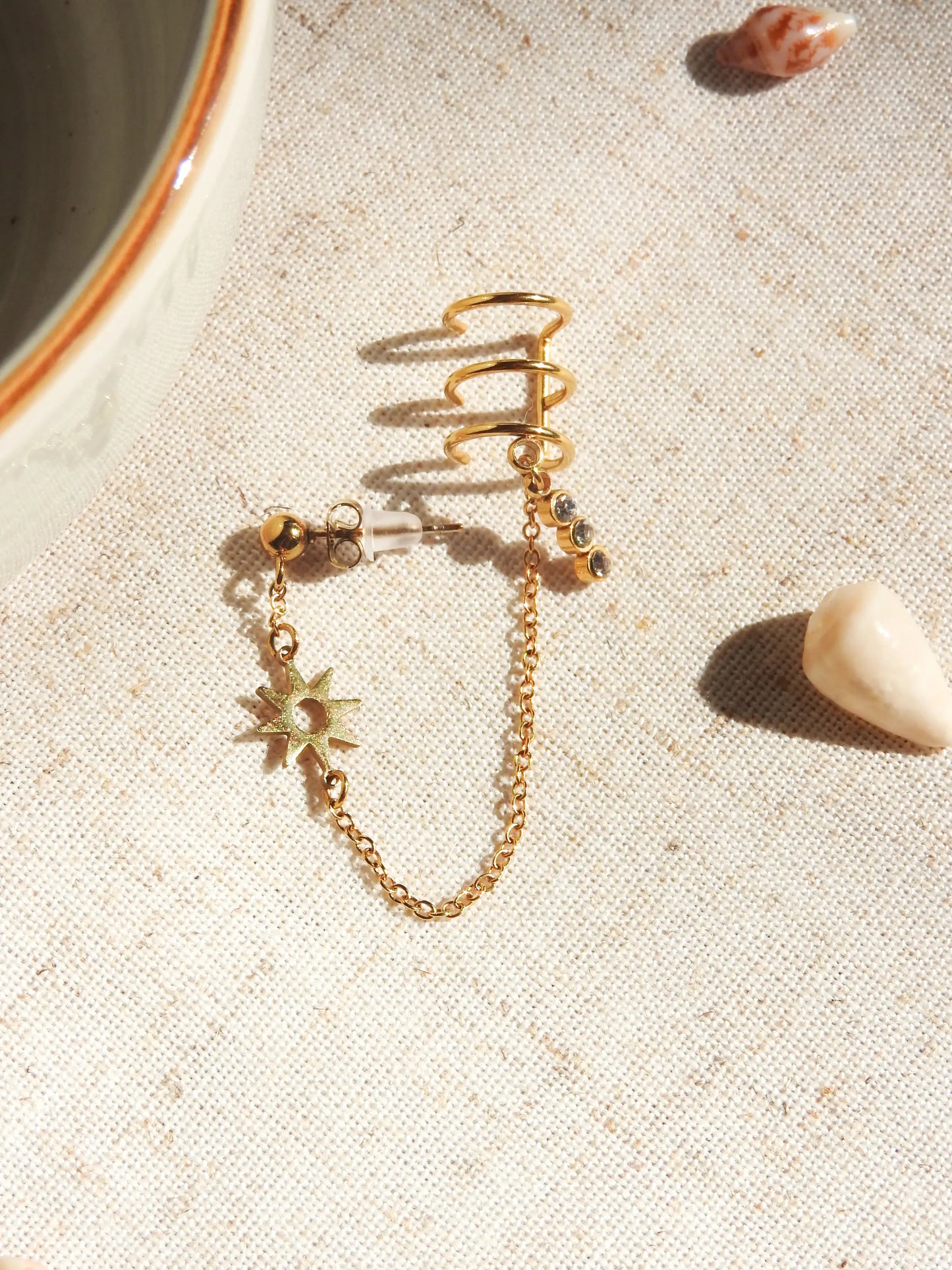 mono-boucle d'oreille clou boule chaînette reliée à une triple bague d'oreille en acier inoxydable doré. Bijou orné d'un soleil en intercalaire et une barre 3 pierres en oxydes de zirconium. Bijou posé à plat sur une nappe beige décorée de coquillages