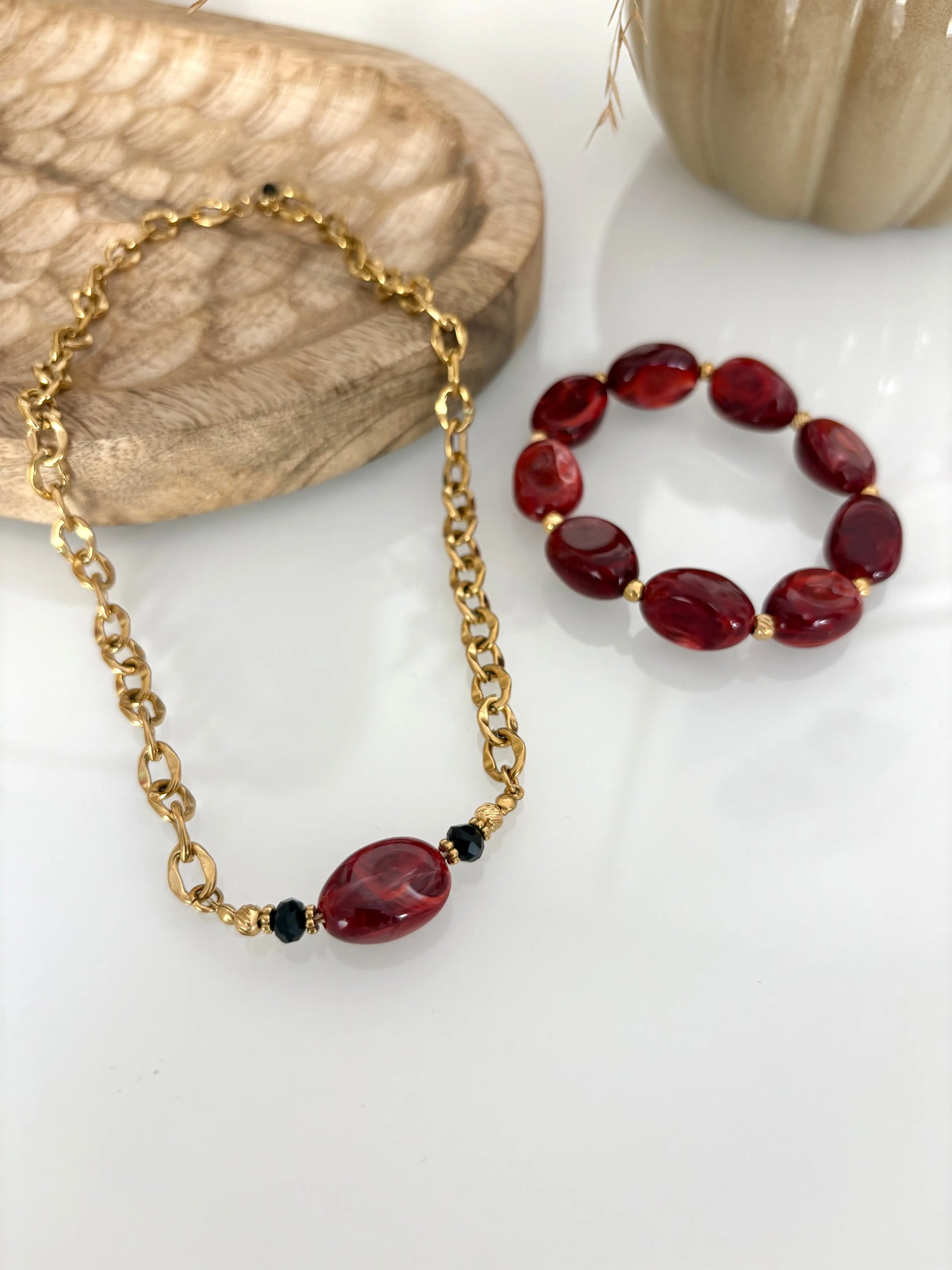 parure de bijoux présentée sur une table en verre. Collier chaîne grosses mailles en acier inoxydable doré, orné d'une grande perle irrégulière bordeaux. Bracelet à grosses perles bordeaux. Table décorée d'un plateau en bambou et d'une citrouille en céramique kaki.