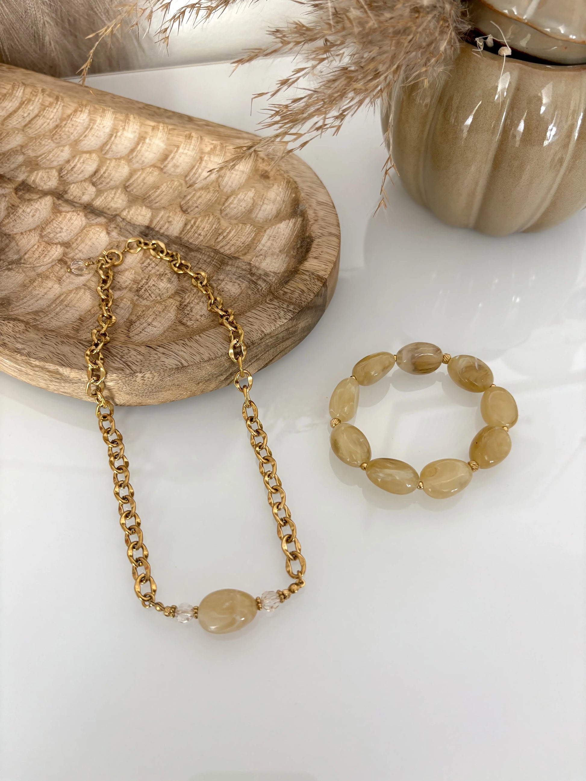 parure de bijoux présentée sur une table en verre. Collier chaîne grosses mailles en acier inoxydable doré, orné d'une grande perle irrégulière kaki. Bracelet à grosses perles kaki. Table décorée d'un plateau en bambou et d'une citrouille en céramique kaki.