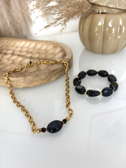 parure de bijoux présentée sur une table en verre. Collier chaîne grosses mailles en acier inoxydable doré, orné d'une grande perle irrégulière noire. Bracelet à grosses perles noires. Table décorée d'un plateau en bambou et d'une citrouille en céramique kaki.