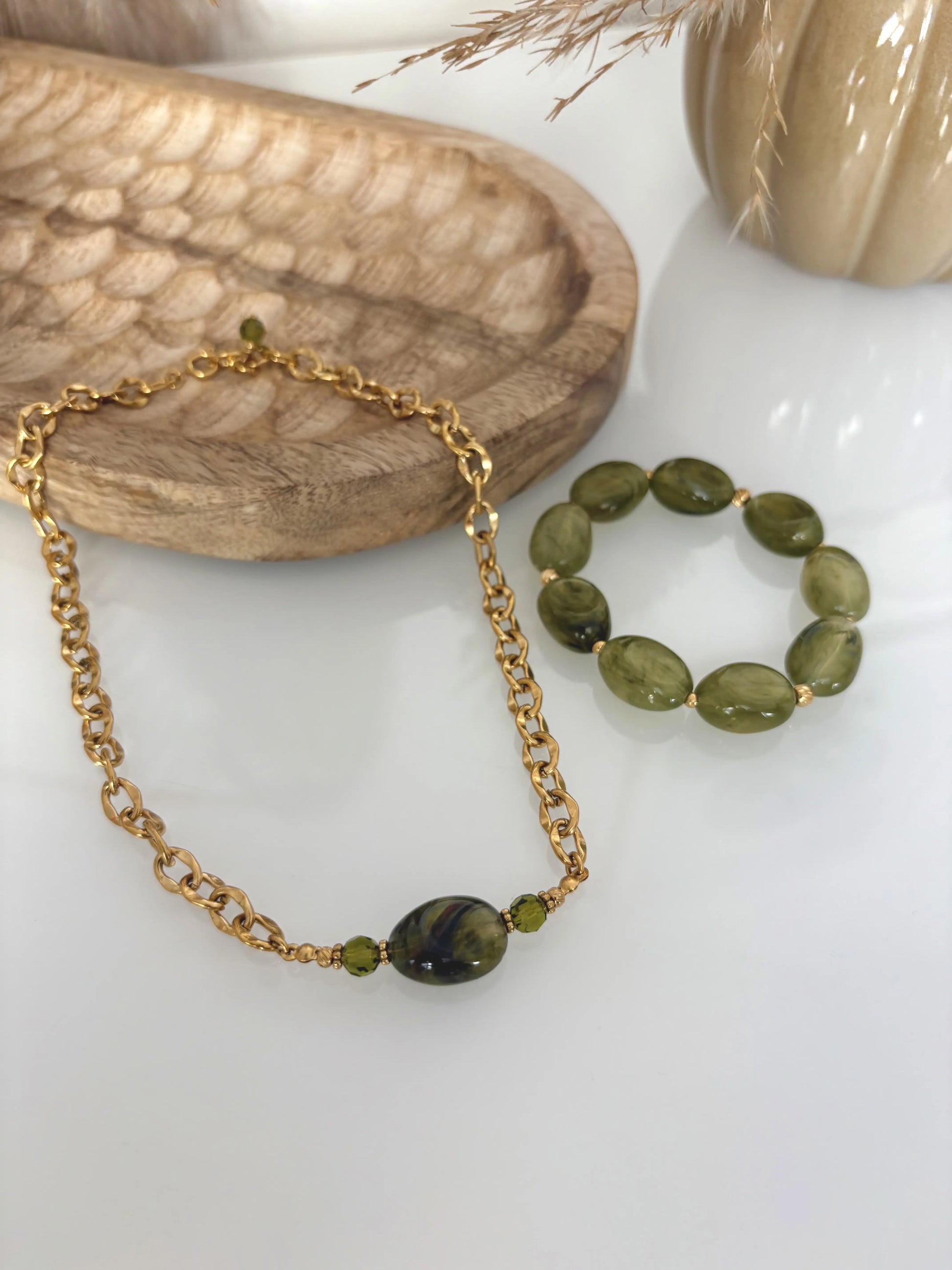 parure de bijoux présentée sur une table en verre. Collier chaîne grosses mailles en acier inoxydable doré, orné d'une grande perle irrégulière vert olive. Bracelet à grosses perles vert olive. Table décorée d'un plateau en bambou et d'une citrouille en céramique kaki.