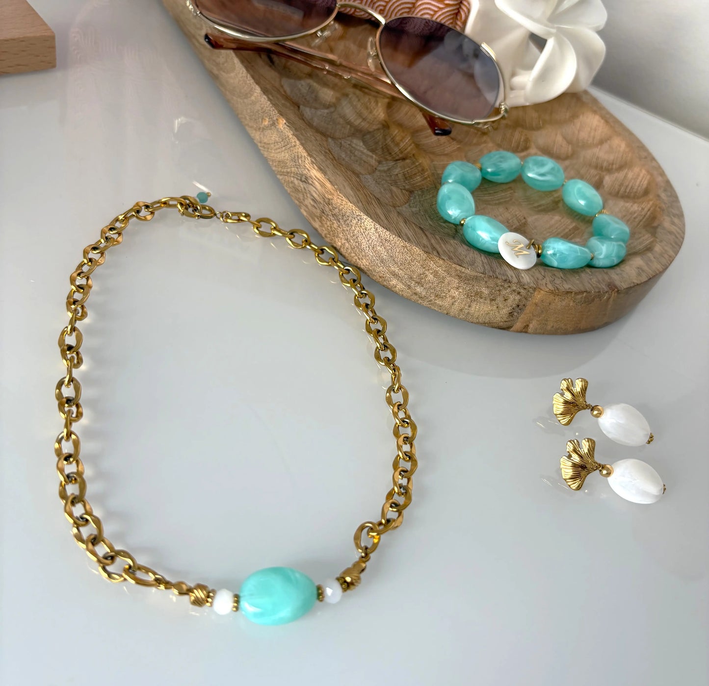parure de bijoux disposés sur une table décorée d'un plateau en bambou contenant des lunettes, une pince fleur et un bracelet de perles turquoise. Collier ras-du-cou a chaîne en acier inoxydable doré et orné d'une perle turquoise et des boucles d'oreilles assorties avec des clous ginkgo et des grandes perles blanches suspendues.