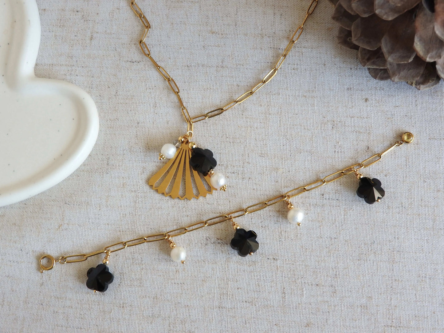 Parure de bijoux posée à plat sur une nappe beige, composé d'un collier chaîne mailles rectangle et pendentif éventail en acier inoxydable doré et perles d'eau douce et perle en forme de fleur noire. Bracelet chaine maille rectangle en acier inoxydable doré, orné de breloques en perles d'eau douce blanche et perles fleurs en verre noire.