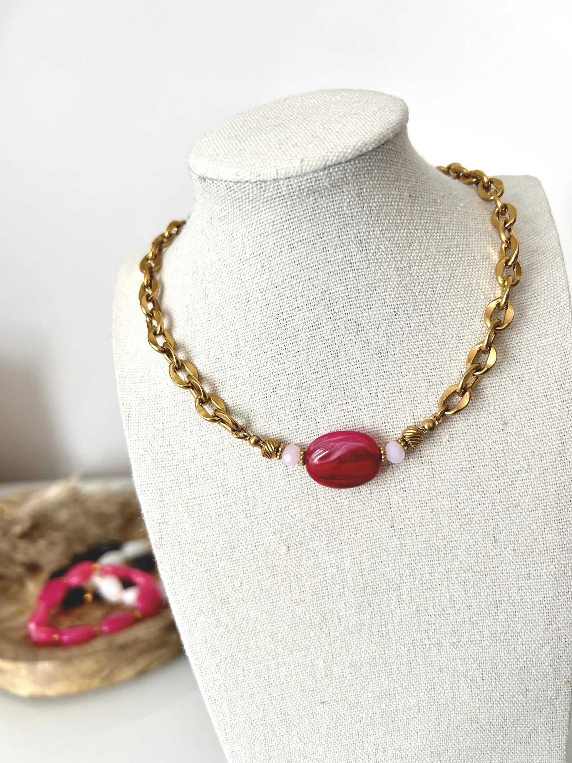 collier à chaîne grosses mailles en acier inoxydable doré. Bijou orné d'une grande perle irrégulière plate de couleur rose fuchsia, posé sur un présentoir buste.