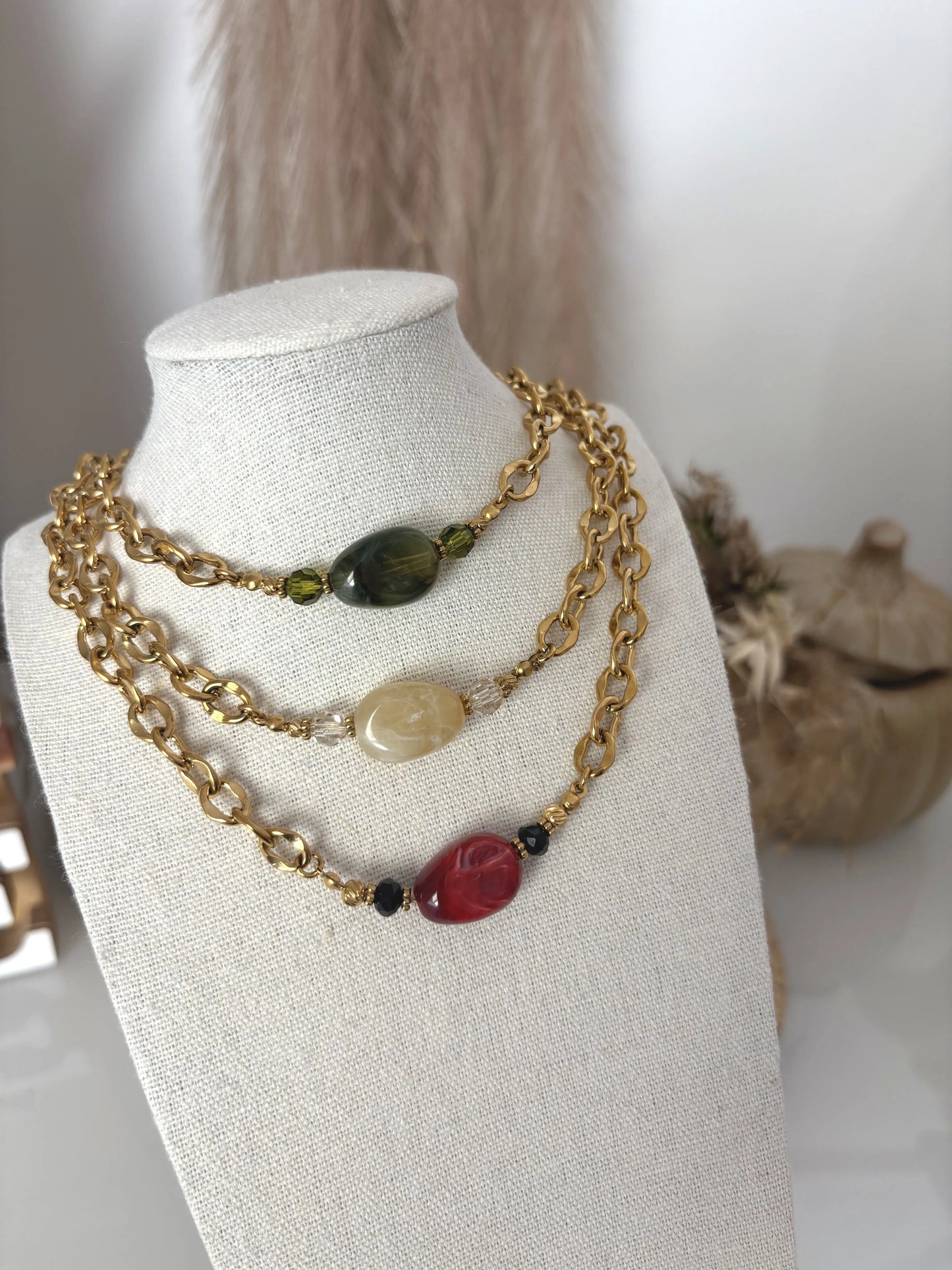 Colliers choker présentés sur un buste présentoir beige en coton. Colliers chaînes à grosses mailles en acier inoxydable doré et ornés d'une grande perle plate irrégulière colorée : vert olive, kaki  et bordeaux.