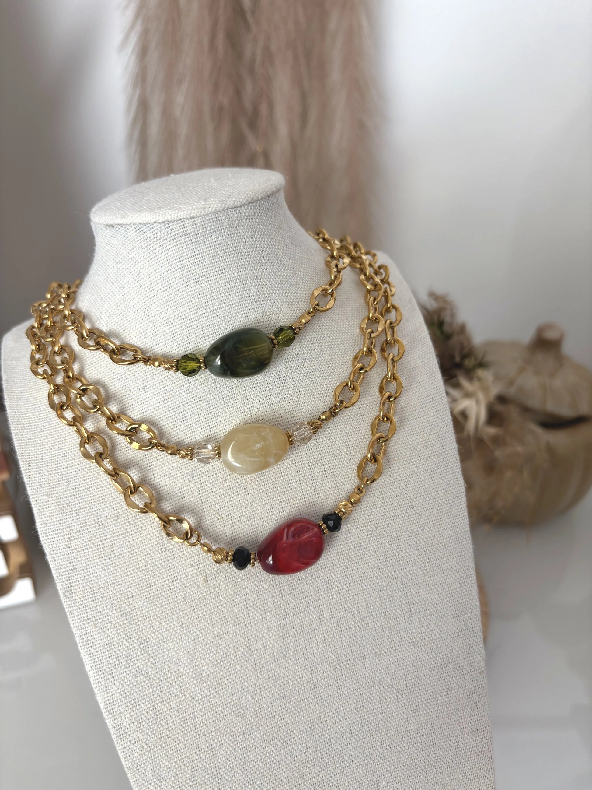 Colliers choker présentés sur un buste présentoir beige en coton. Colliers chaînes à grosses mailles en acier inoxydable doré et ornés d'une grande perle plate irrégulière colorée : vert olive, kaki  et bordeaux.