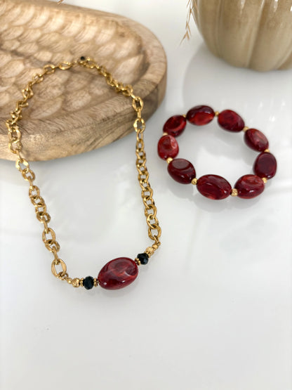 Collier choker à grosses mailles en acier inoxydable doré et orné d'une perle irrégulière bordeaux. Bijou présenté sur une table en verre trempée et décorée d'un plateau en bambou et un bracelet de perles bordeaux.