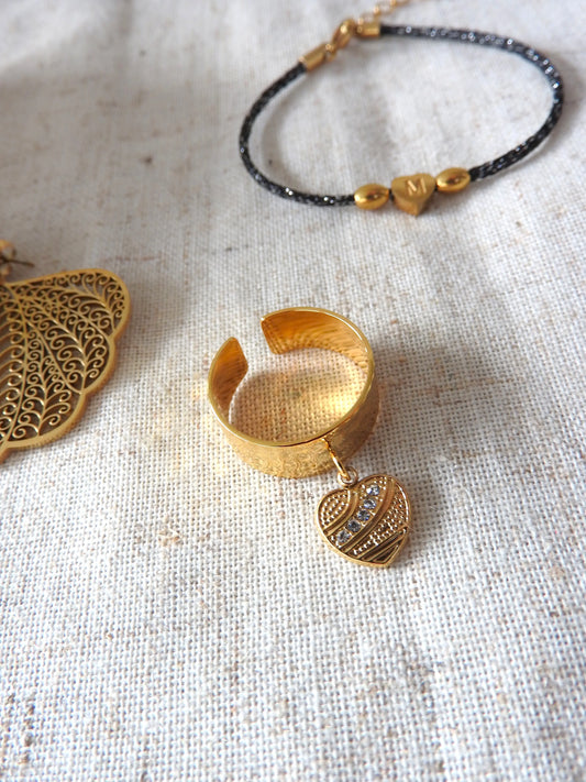 Bague à anneau martelé réglable en acier inoxydable doré ornée d'une breloque coeur avec des pierres en oxyde de zirconium. Bijou posé sur une nappe beige en coton et posé à côté d'un bracelet cordon noir à fil argenté et orné de perles en acier doré dont une perle coeur à lettre M gravée.