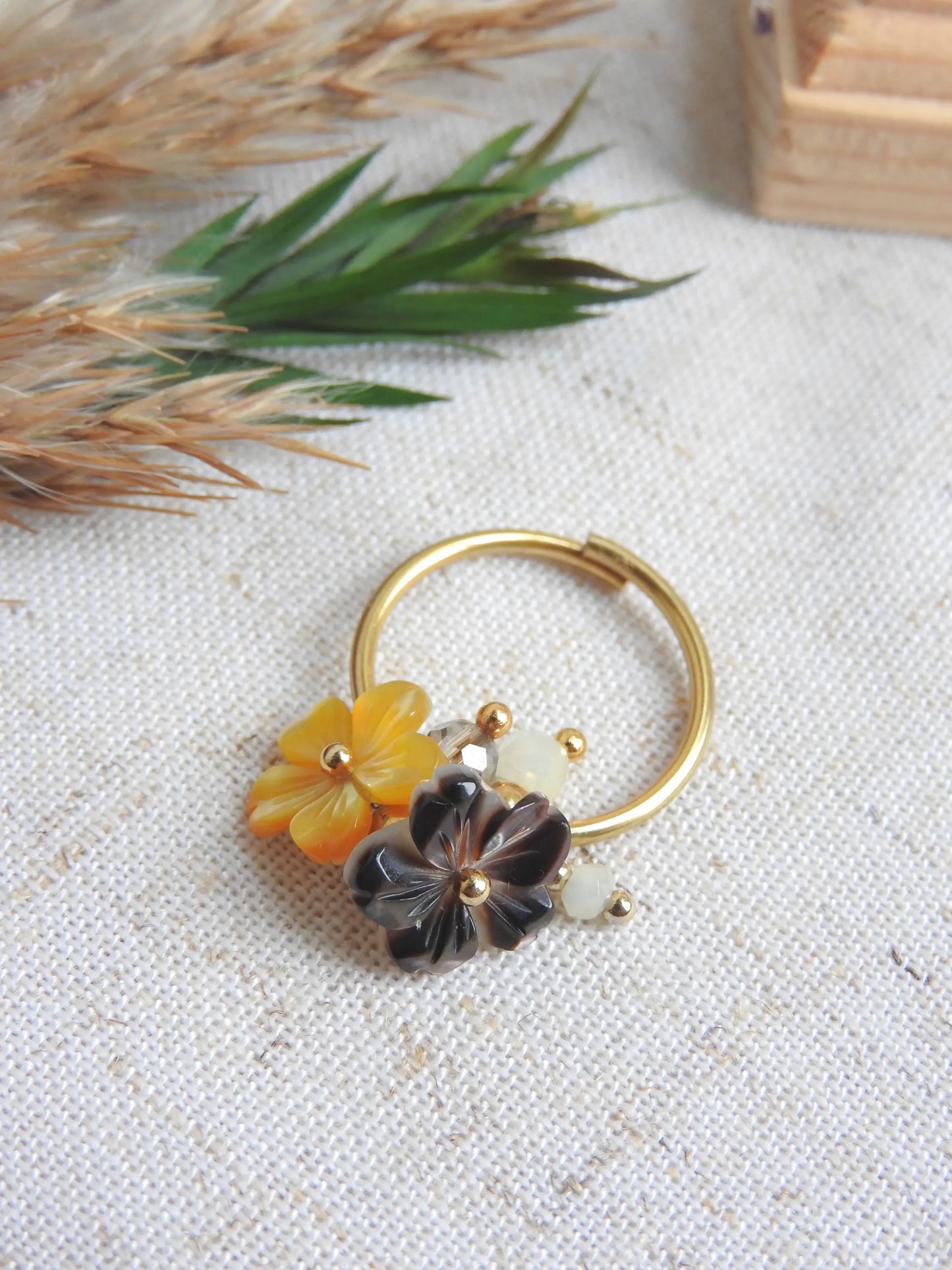 bague à anneau fin réglable en acier inoxydable doré posé à plat sur une nappe beige, décorée d'un mini bouquet de fleurs. Bijou orné de breloques fleurs en nacre jaune et marron et de perles de verre et en acier inoxydable doré.