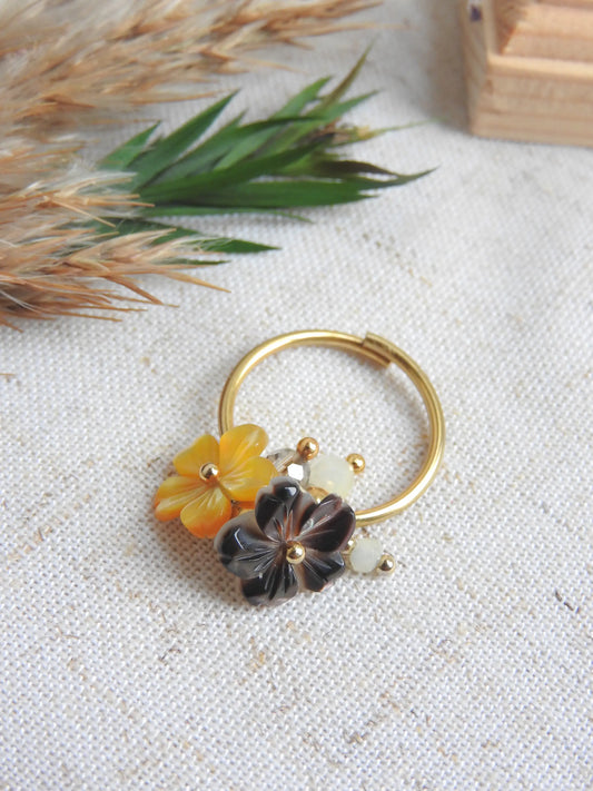 bague à anneau fin réglable en acier inoxydable doré posé à plat sur une nappe beige, décorée d'un mini bouquet de fleurs. Bijou orné de breloques fleurs en nacre jaune et marron et de perles de verre et en acier inoxydable doré.