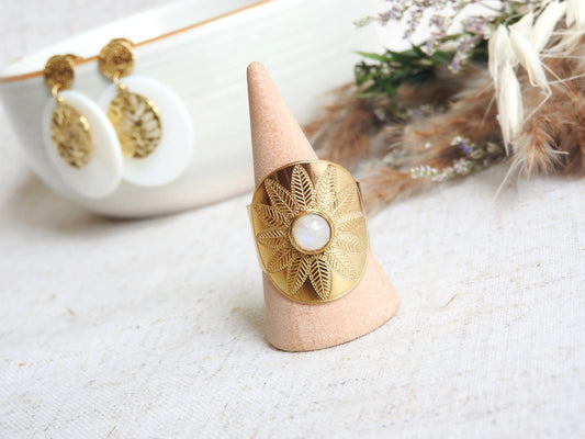 bague style bohème, ethnique réglable en acier inoxydable doré, ornée d'une pierre de lune. Bijou posé sur un cône en bambou posé sur une nappe beige, avec en fond des boucles d'oreilles sequin en nacre blanche suspendues à un bol en céramique.