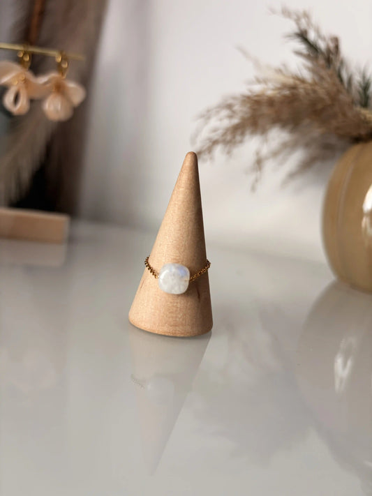 Bague chaînette en acier inoxydable doré et ornée d'une pierre de lune carré. Bijou présenté sur un cône cylindrique en bambou.