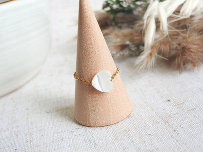 bague chaînette en acier inoxydable doré ornée d'une perle coeur en nacre blanche. Bijou posé sur un cône en bambou posé sur une nappe beige décorée d'un mini-bouquet de fleurs.