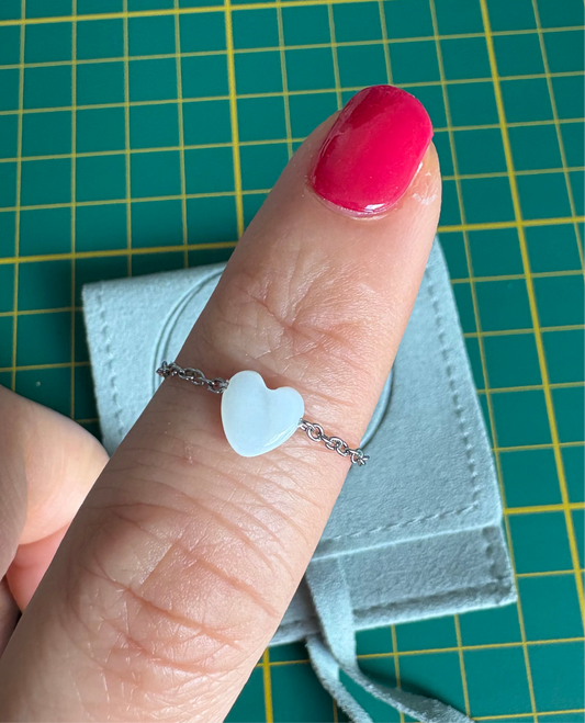 bague chaînette en acier inoxydable argenté et ornée d'une perle coeur en nacre blanche. Bijou porté à l'index avec en fond un pochon en micro-fibre.