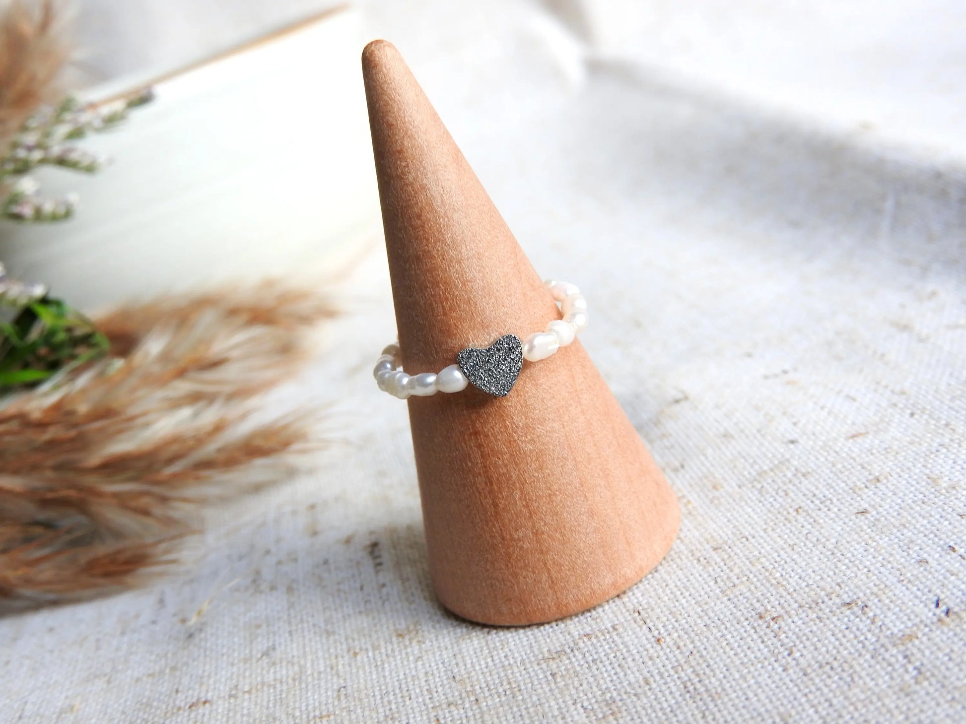 Bague élastique en perles d'eau douce blanche et perle cœur à effet diamanté en acier inoxydable argenté. Bijou posé sur un cône en bambou. 
