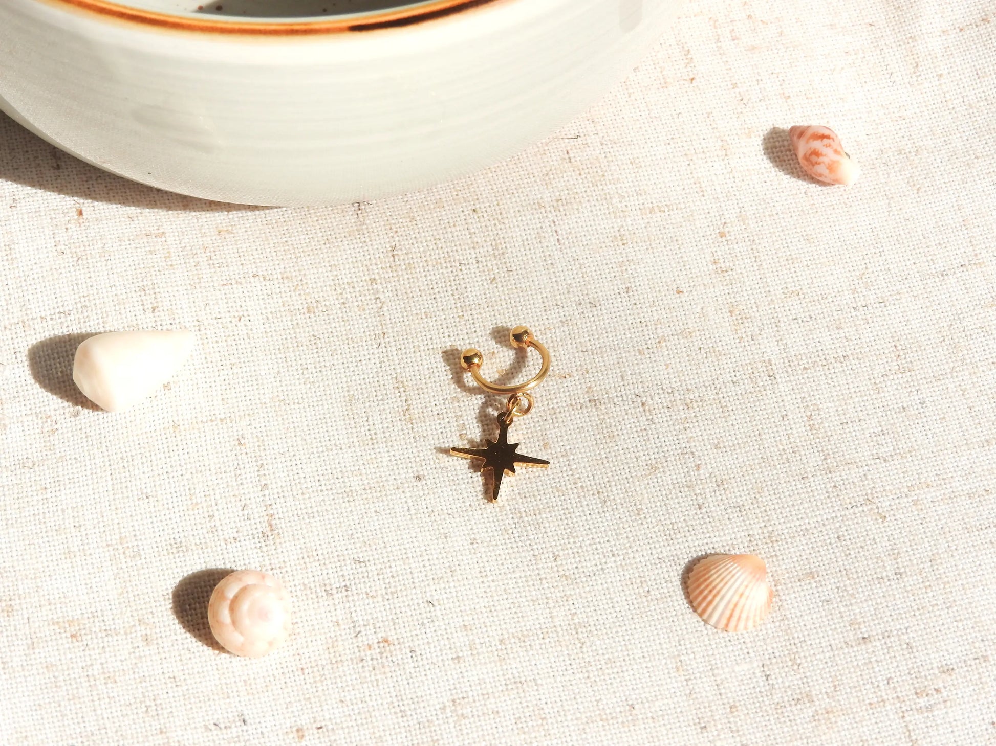 bague d'oreille à boules dévissables en acier inoxydable doré avec un charm etoile du nord. Bijou posé à plat sur une nappe beige décorée de coquillages.