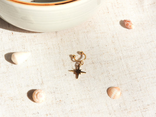 bague d'oreille à boules dévissables en acier inoxydable doré avec un charm etoile du nord. Bijou posé à plat sur une nappe beige décorée de coquillages.