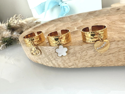 bagues à anneau réglable breloques en acier inoxydable doré, disposées sur un plateau en bambou sur une table en verre décorée d'un mini-bouquet de fleurs. Breloque fleur en nacre blanche, breloque coeur ornée de pierre en oxydes dezirconium et breloque coeur papillon strass.