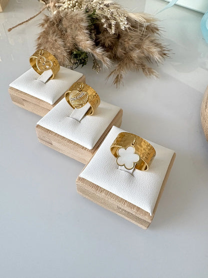 bagues à anneau réglable breloques en acier inoxydable doré, disposées sur des supports individuels en bambou sur une table en verre décorée d'un mini-bouquet de fleurs. Breloque fleur en nacre blanche, breloque coeur ornée de pierre en oxydes dezirconium et breloque coeur papillon strass.