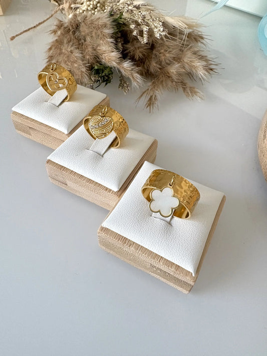 bagues à anneau réglable breloques en acier inoxydable doré, disposées sur des supports individuels en bambou sur une table en verre décorée d'un mini-bouquet de fleurs. Breloque fleur en nacre blanche, breloque coeur ornée de pierre en oxydes dezirconium et breloque coeur papillon strass.