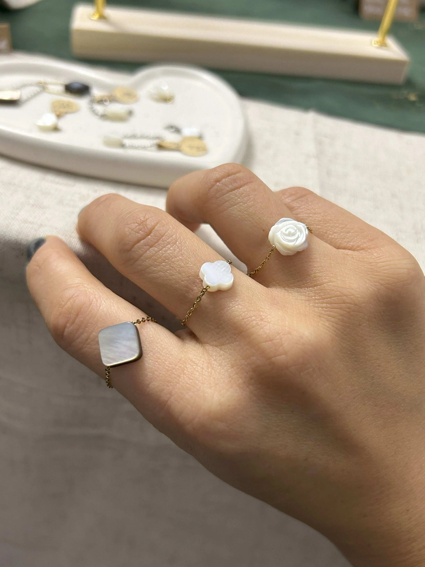 trois bagues chaînettes en acier inoxydable doré portées aux doigts d'une main. A l'index, une perle carré aux reflets noir et gris, au majeur une perle trèfle en nacre blanche et à l'annulaire une perle rose en nacre blanche. 