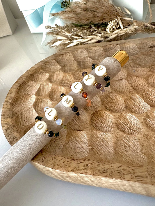 Bagues perles de verre colorées et acier inoxydable doré, ornées d'une perle plate en nacre blanche avec motif lettre initiale dorée. Bijoux posés et alignés sur une boudin en tissu beige, posé sur une plateau en bambou.