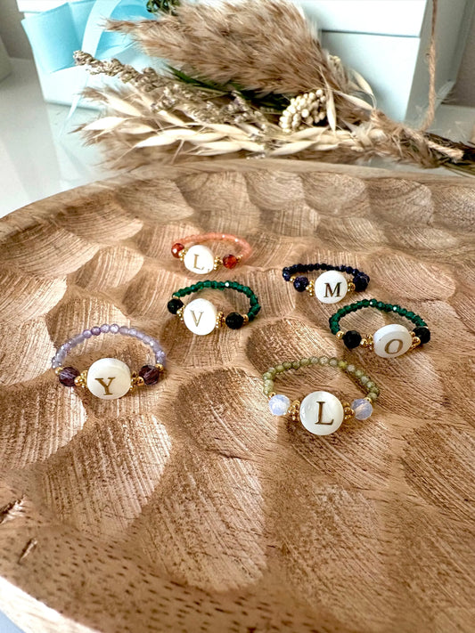 Bagues perles de verre colorées et acier inoxydable doré, ornées d'une perle plate en nacre blanche avec motif lettre initiale dorée. Bijoux posés sur une plateau en bambou.