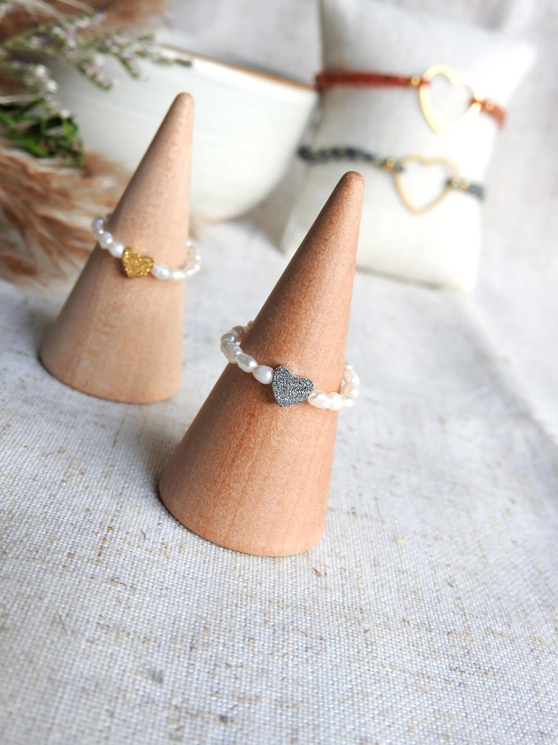 Bagues élastique en perles d'eau douce blanche et perle coeur à effet diamanté en acier inoxydable doré en avant-plan et doré en arrière-plan. Bijoux posés sur un cône en bambou. 
