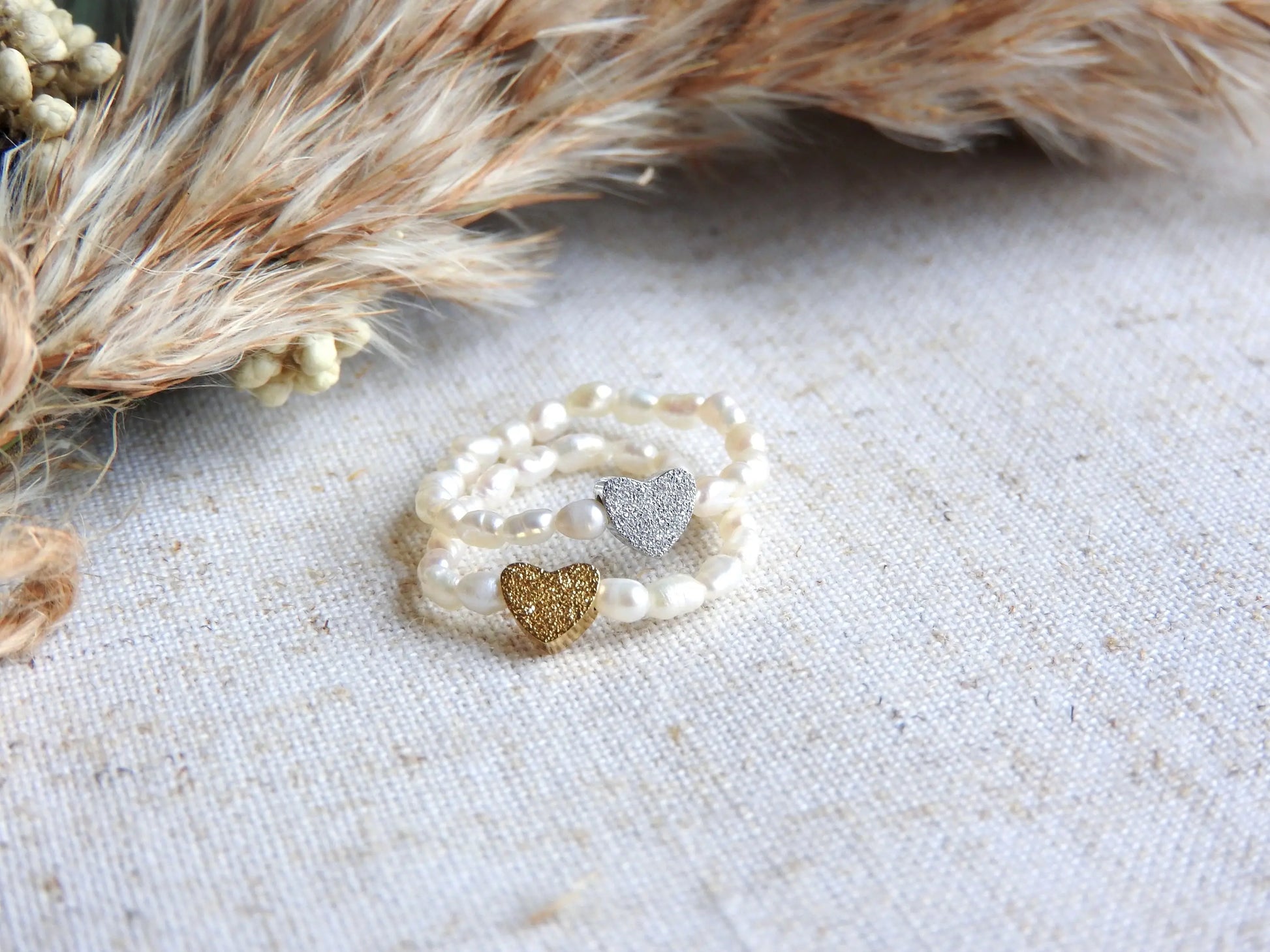 Bagues élastique en perles d'eau douce blanche et perle cœur à effet diamanté en acier inoxydable doré et argenté. Bijoux superposés sur une nappe beige en coton, décorée de brindilles.