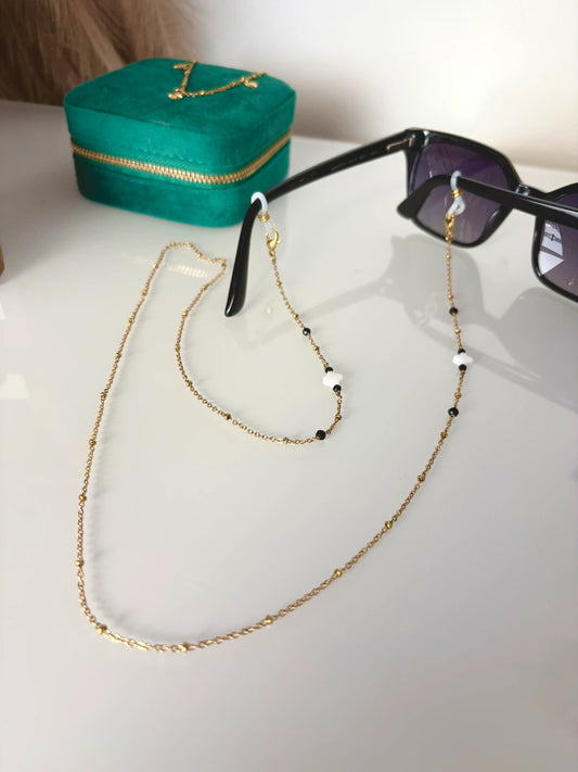 Bijou chaîne de lunettes en acier inoxydable doré et perlée, ornée de perles en nacre blanche et perles de verre noir. Bijou monté sur une monture de lunettes de soleil noire.