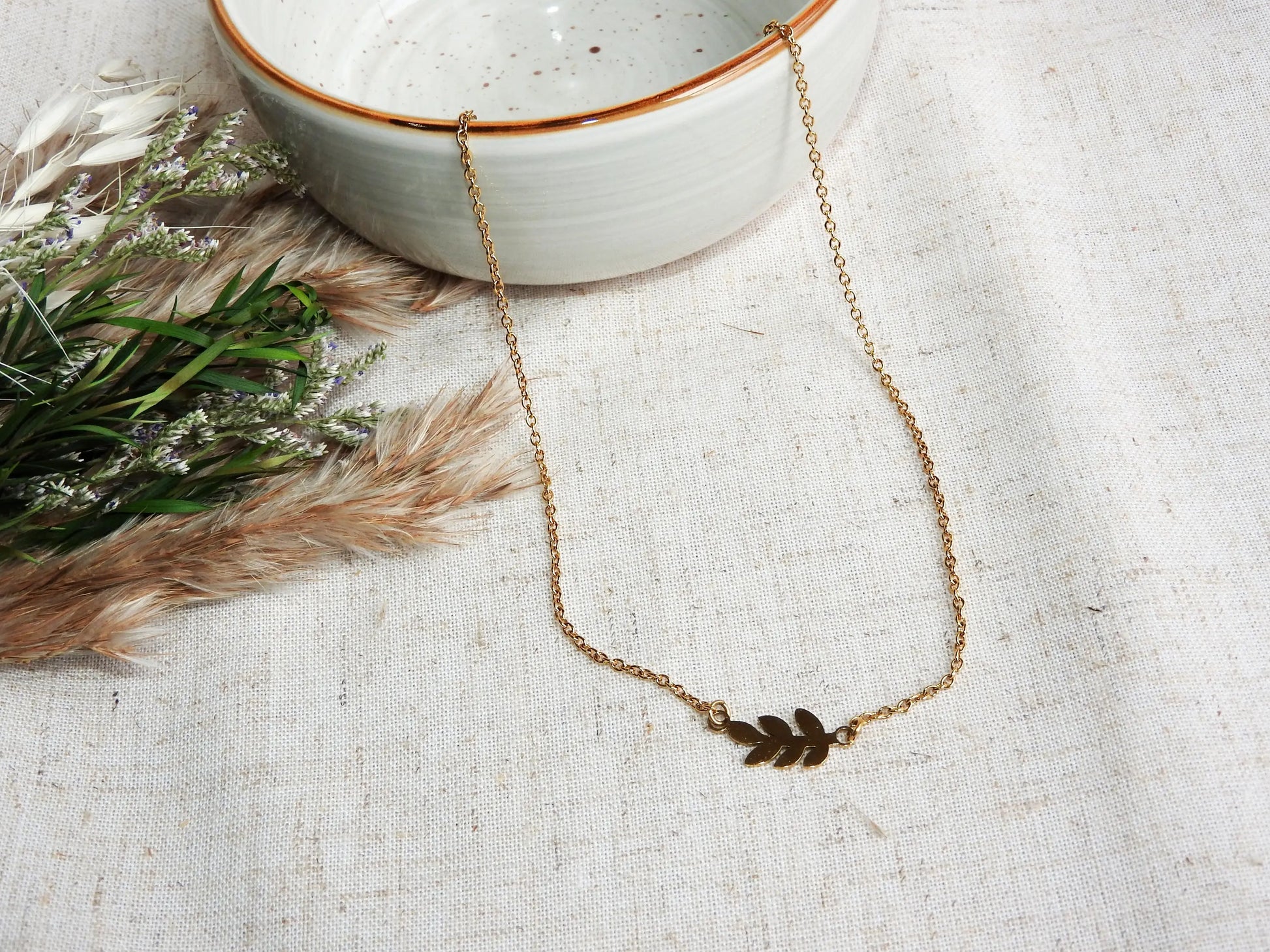 Collier à chaîne fine orné d'une feuille intercalaire en acier inoxydable doré. Bijou posé sur une nappe beige et un bol en céramique vert pâle et décorée d'un mini-bouquet de fleurs.
