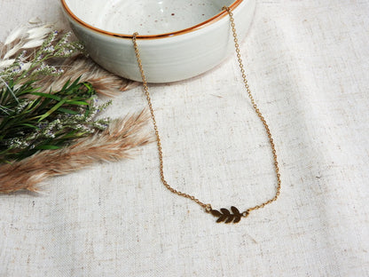 Collier à chaîne fine orné d'une feuille intercalaire en acier inoxydable doré. Bijou posé sur une nappe beige et un bol en céramique vert pâle et décorée d'un mini-bouquet de fleurs.
