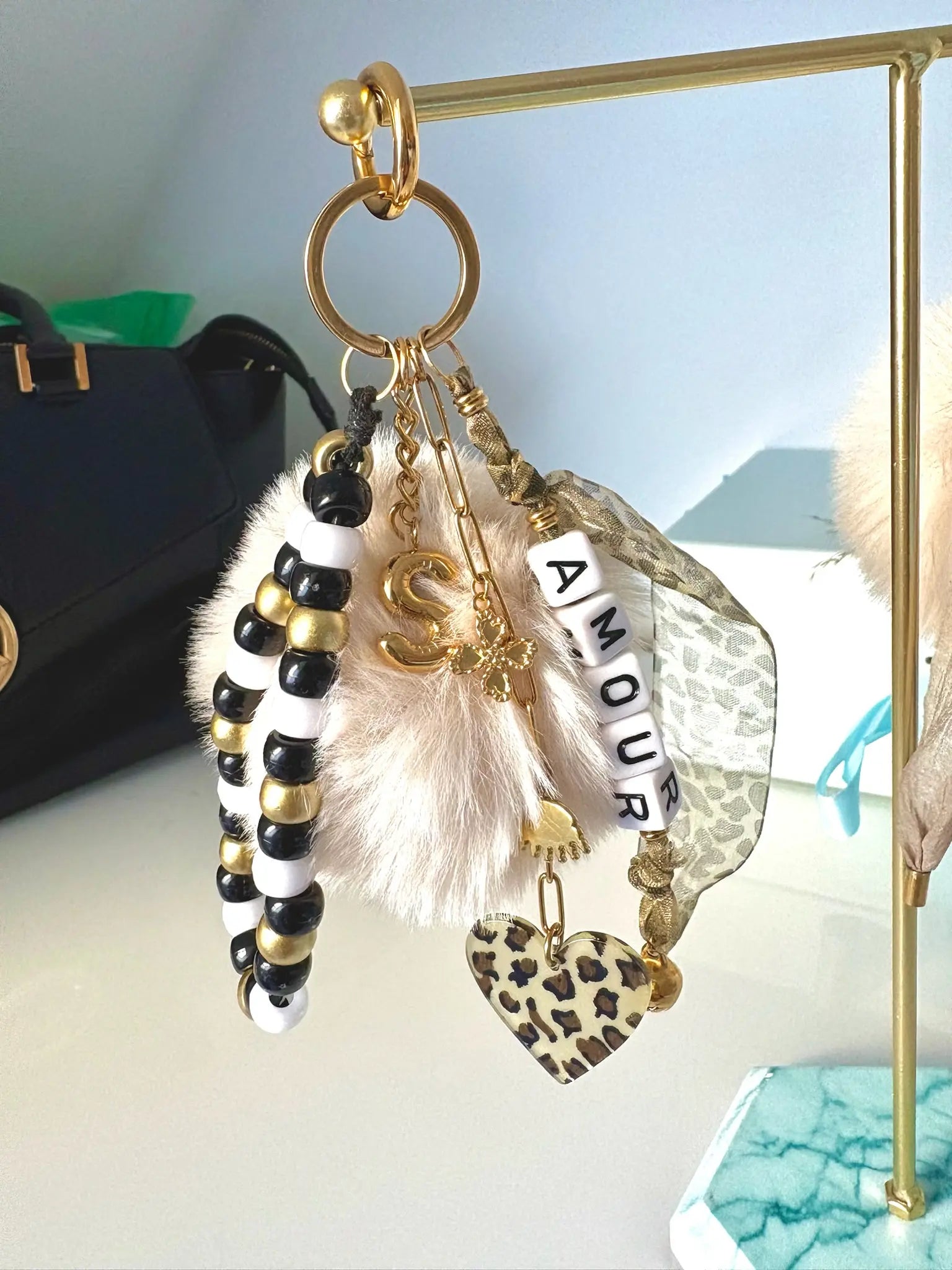 Bijou de sac et porte-clés posés sur un présentoir métallique doré. Accessoire à personnaliser, lettre initiale S effet ballon, ruban motif léopard, cœur motif léopard, bracelet en perles coloris noir, doré et blanc et chaînette en acier inoxydable doré avec breloques trèfle, oeil protecteur et grelot.