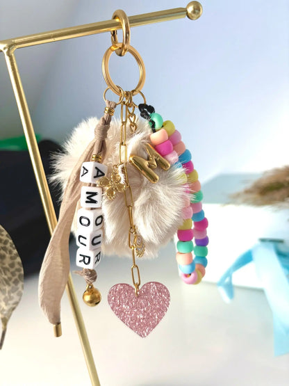 Bijoux de sac et porte-clés posés sur un présentoir métallique doré. Accessoire à personnaliser, lettre initiale M effet ballon, ruban couleur moka, cœur rose paillette, bracelet en perles multicouleurs et chaînette en acier inoxydable doré avec breloques trèfle, oeil protecteur et grelot.