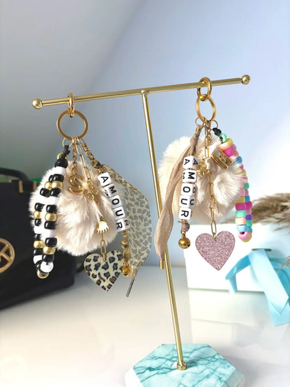 Bijoux de sac et porte-clés posés sur un présentoir métallique doré. Accessoires à personnaliser, lettre initiale S et M à effet ballon, ruban motif léopard et moka, cœurs motif léopard et rose paillette, bracelets en perles coloris noir, doré et blanc pour l'un et l'autre multicouleurs et chaînettes en acier inoxydable doré avec breloques trèfle, oeil protecteur et grelot.