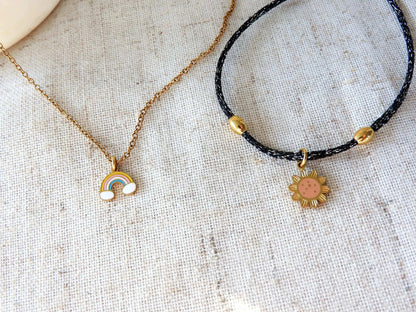 Collier enfant à chaîne fine en acier inoxydable doré et orné d'un pendentif arc-en-ciel coloré en résine époxy et acier inoxydable doré et posé à côté d'un bracelet cordon noir scintillant orné d'un pendentif tournesol en résine époxy et acier inoxydable doré. 