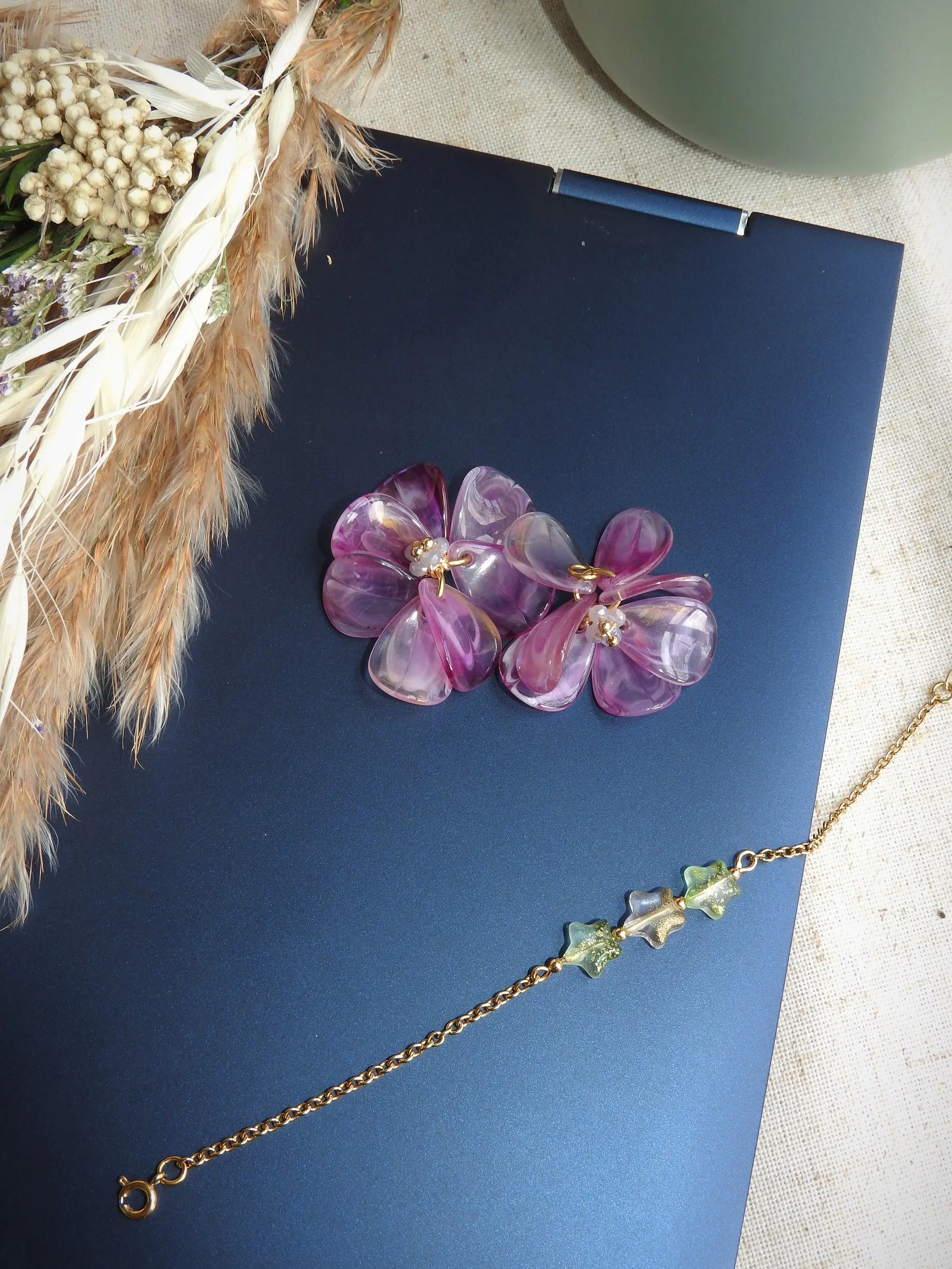 Suspensions pour créoles en acier inoxydable assemblées à la main. Bouquet floral réalisé à partir de pétales en acrylique violet marbré et de perles de verre colorées et en acier inoxydable doré. Bijou posé sur le dessus d'un ordinateur portable avec un bracelet chaîne orné de perles en forme d'étoile scintillante couleur vert pomme.