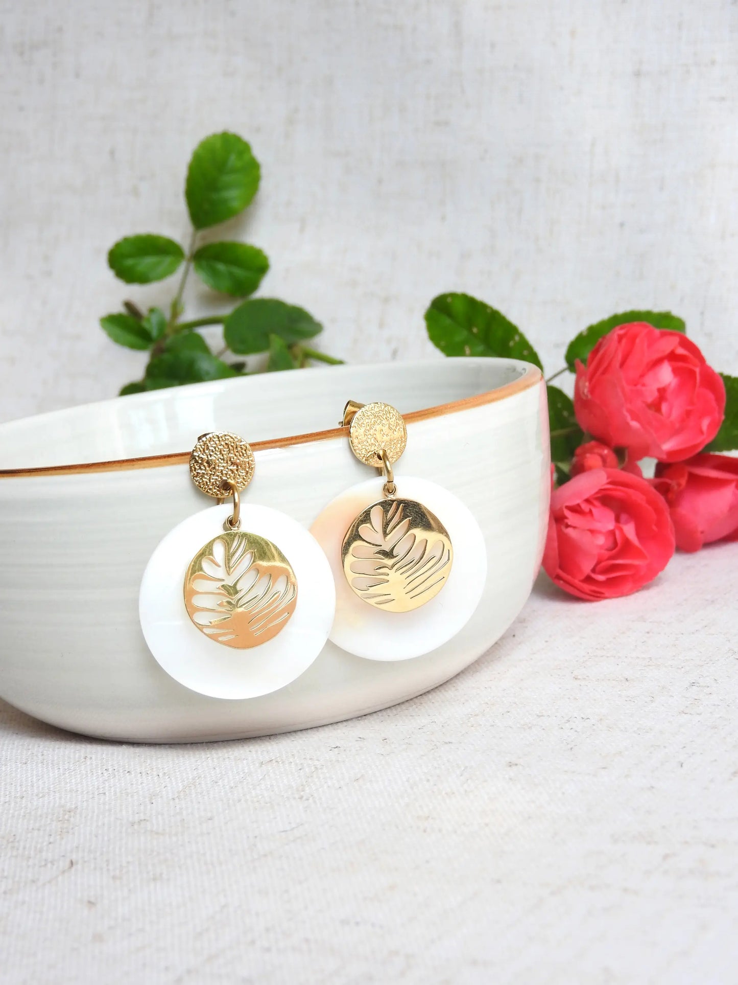 boucles d'oreilles clous à disque martelé en acier doré, ornées d'un double sequin, l'un en nacre blanche et l'autre en médaille rond avec une feuille évidée. Bijou suspendu à un bol en céramique posé sur une nappe beige décorée de roses roses. 
