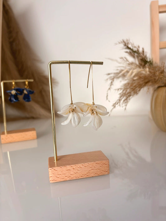 Boucles d'oreilles hameçon en acier inoxydable doré, ornées de pétales de fleurs en acrylique ivoire et perles de verre pour pistil. Bijou présenté sur un support métallique doré et bambou posé sur une table en verre et décorée d'une citrouille décorative en céramique.