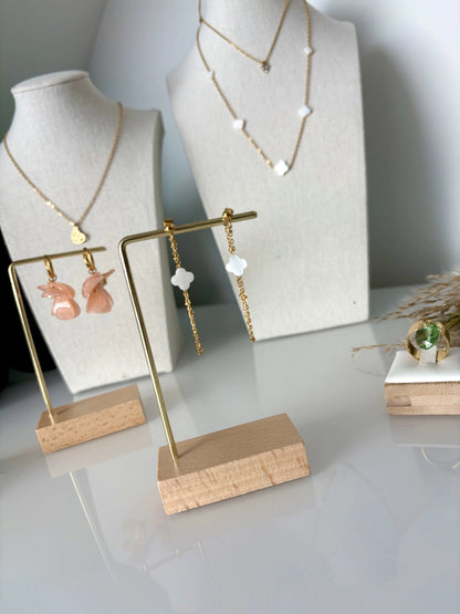 boucles d'oreilles chaînettes en acier inoxydable doré ornées d'une perle trèfle en nacre blanche. Bijou suspendu à un présentoir en métal doré et bambou. En arrière plan, deux présentoirs bijoux buste présentant des colliers en acier doré à pendentif coeur papillon, un autre avec 3 pierres en zircon et un autre avec des perles trèfle en nacre blanche intercalées. D'autres boucles d'oreilles sur le présentoir à boucle d'oreilles en pétales de couleur pêche en acrylique montées sur des mini-créoles en acier.