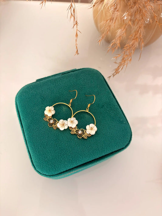 Boucles d'oreilles crochet en acier inoxydable doré et ornées de strass et petites fleurs en nacre blanche. Bijoux posés sur une boite à bijoux vert emeraude.