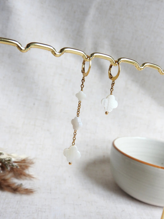 boucles d'oreilles à chaînes pendantes asymétriques ornées de perles trèfle en nacre blanche de tailles variées. Bijou suspendues sur une barre métallique dorée. 