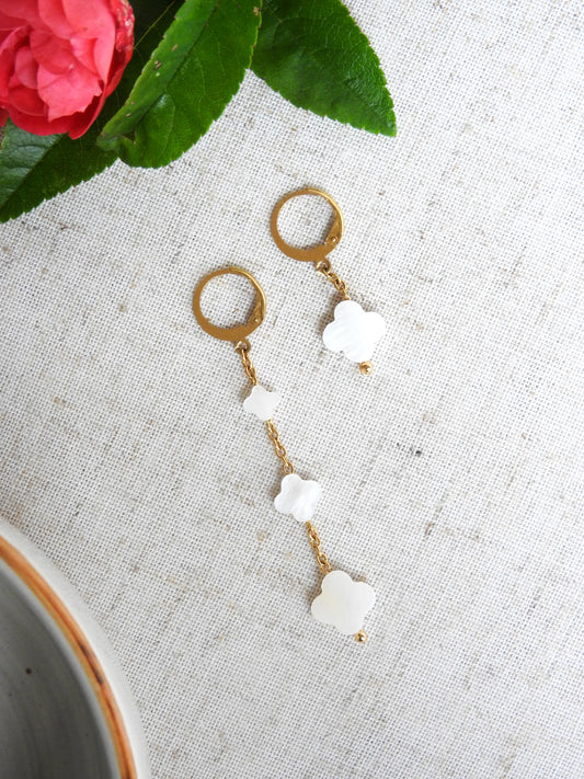 boucles d'oreilles à chaînes pendantes asymétriques ornées de perles trèfle en nacre blanche. Bijou posé à plat sur une nappe blanche décorée de roses. 