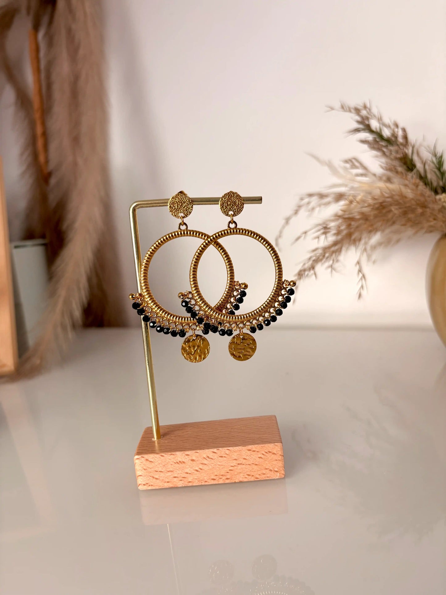 boucles d'oreilles chandelier en acier inoxydable doré disposées sur un support métallique doré et bambou. Bijoux ornés de perles de verre noires et sequins ronds martelés.