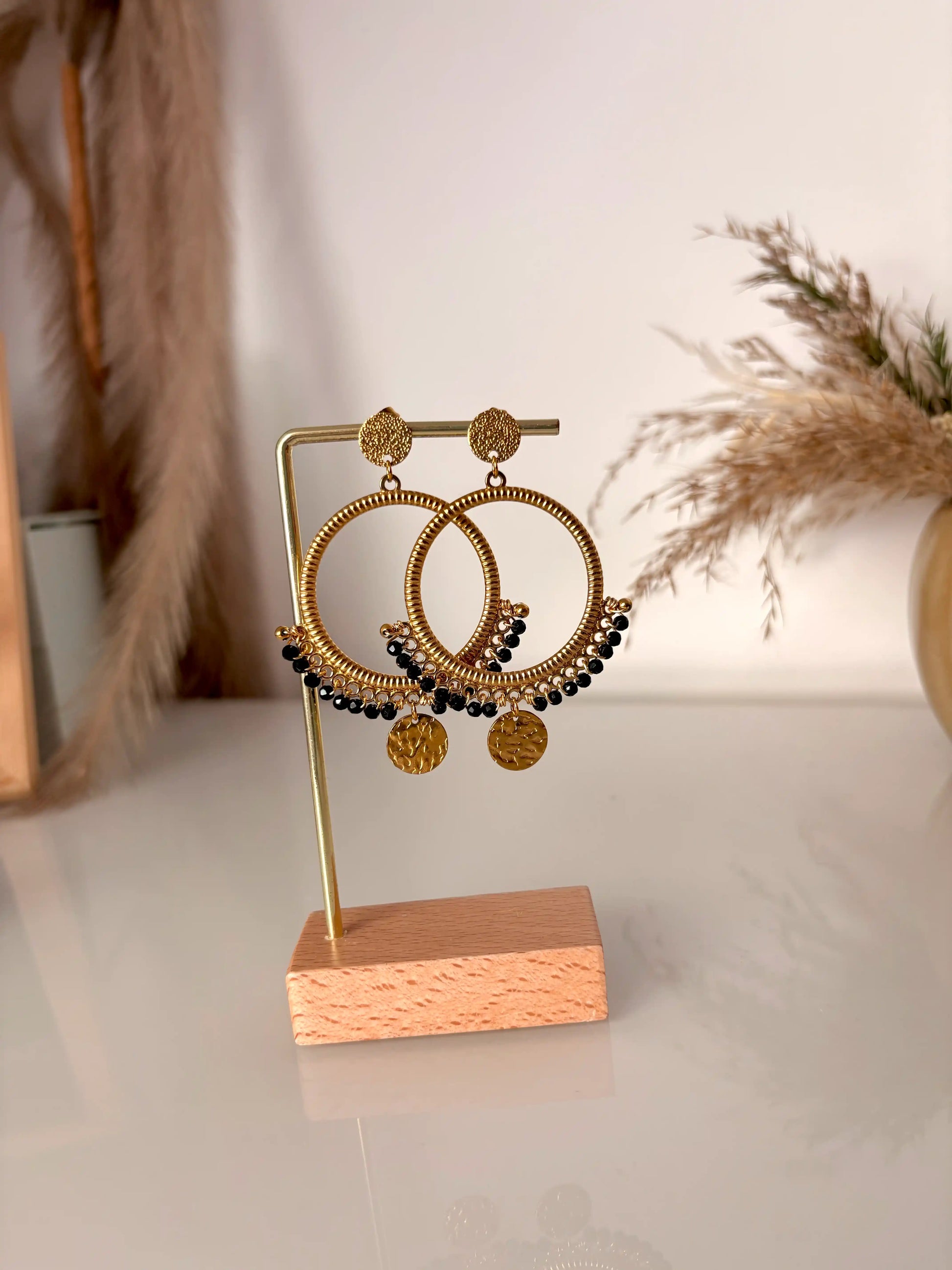boucles d'oreilles chandelier en acier inoxydable doré disposées sur un support métallique doré et bambou. Bijoux ornés de perles de verre noires et sequins ronds martelés.