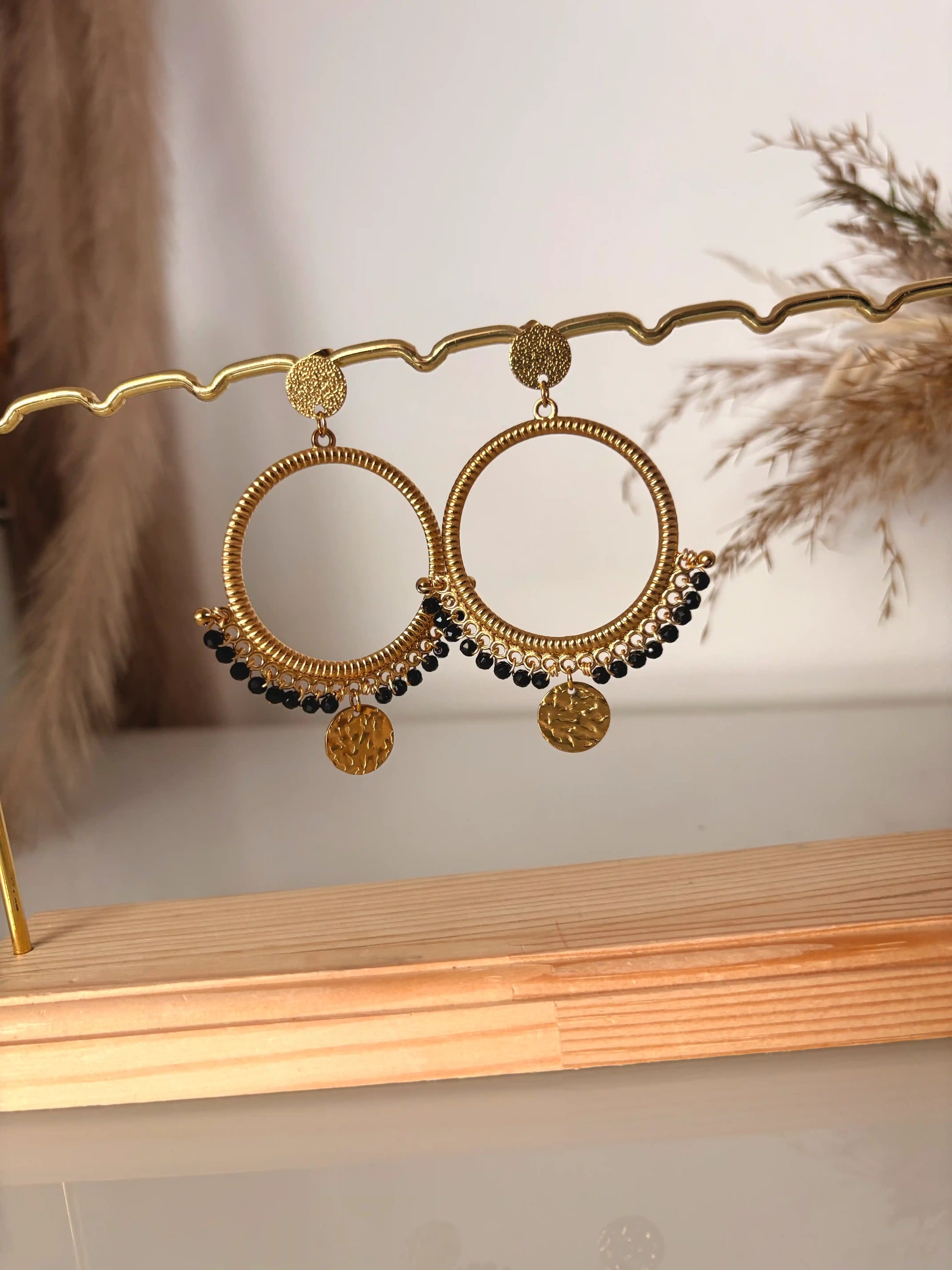 boucles d'oreilles chandelier en acier inoxydable doré et orné de perles noires et de sequins rond martelé. Bijoux présentés sur un support métallique doré et bambou.
