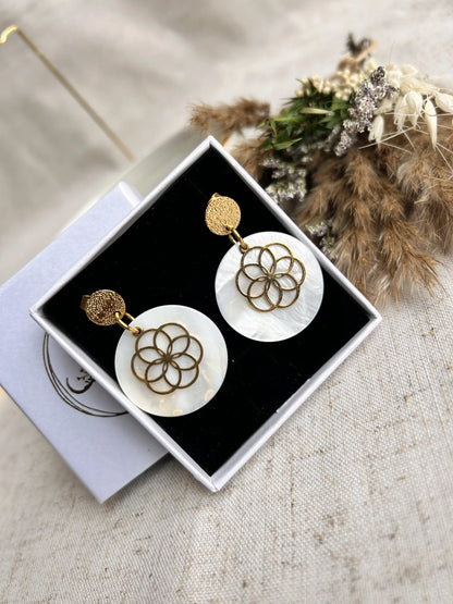 Boucles d'oreilles clous à disque martelé rangées dans un écrin blanc avec une mousse noire. Bijou orné d'un sequin en nacre blanche superposée d'une fleur évidée en acier inoxydable doré.