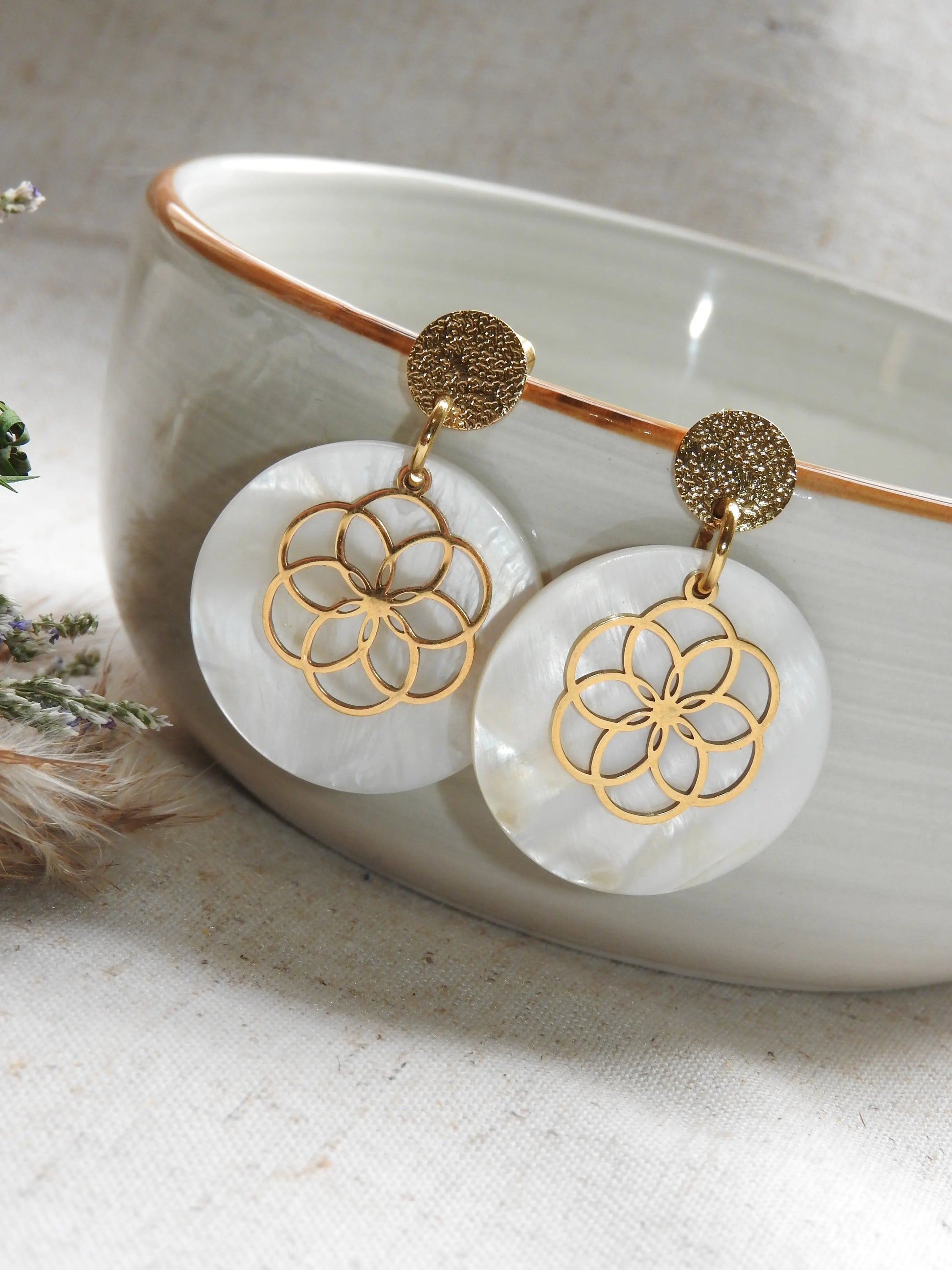 Boucles d'oreilles clous à disque martelé suspendues sur le bord d'un bol en céramique vert pâle. Bijou orné d'un sequin en nacre blanche superposée d'une fleur évidée en acier inoxydable doré.