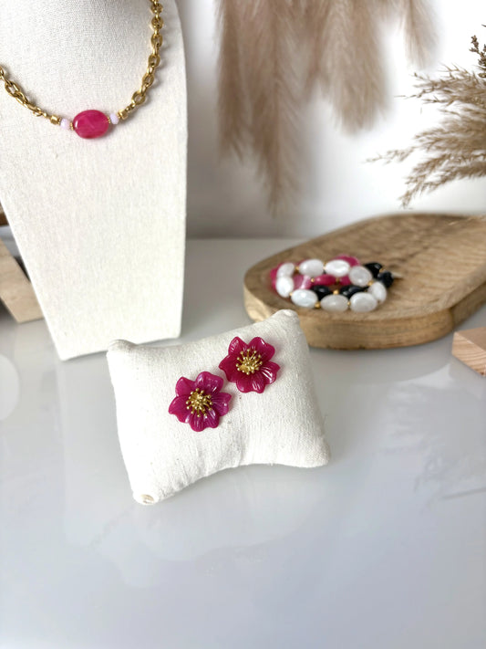 boucles d'oreilles clous à pistil en acier inoxydable doré et orné d'une fleur hibiscus framboise. Bijoux présentés sur un mini-coussin en coton beige, avec en fond un collier à grosse perle ovale fuchsia, posé sur un buste présentoir.