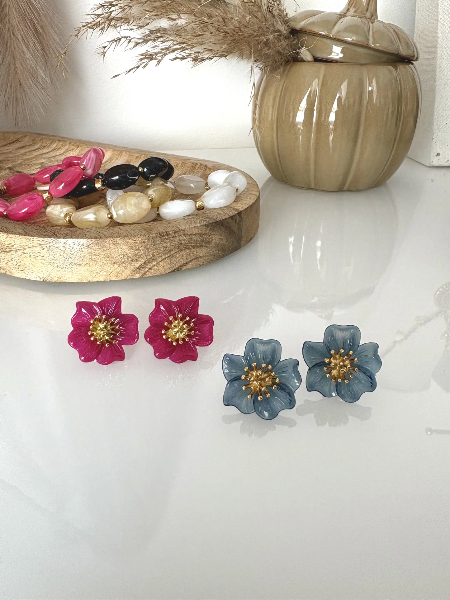 deux paires de boucles d'oreilles clous à pistil en acier inoxydable doré et orné d'une fleur hibiscus bleu foncé et framboise. Bijoux présentés sur une table en verre trempé et décorée d'un bol citrouille kaki avec brindilles et un plateau en bambou où sont disposés des bracelets à perles colorées.